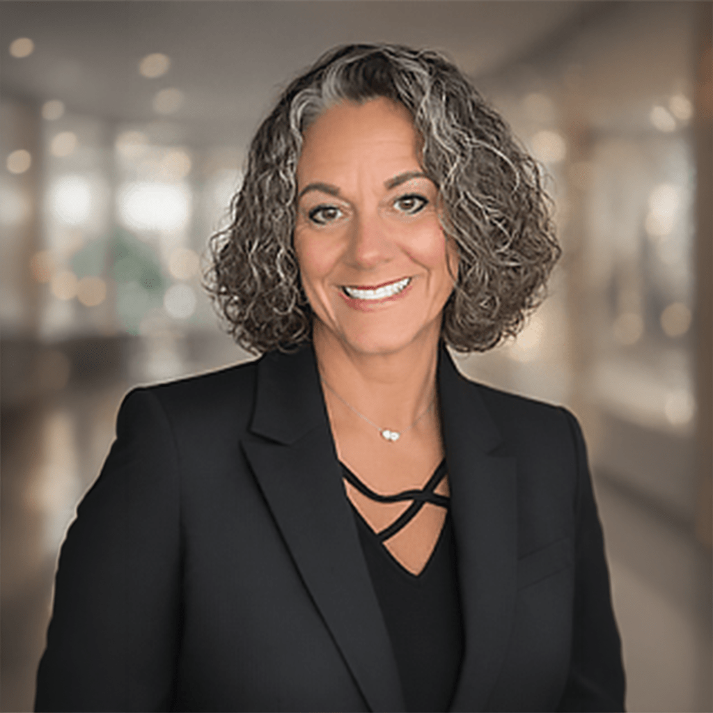 Smiling woman with curly hair in a black blazer, posing against a softly blurred background. Her approachable expression conveys professionalism.