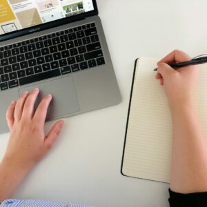 Person is working on a laptop and writing in a notebook.